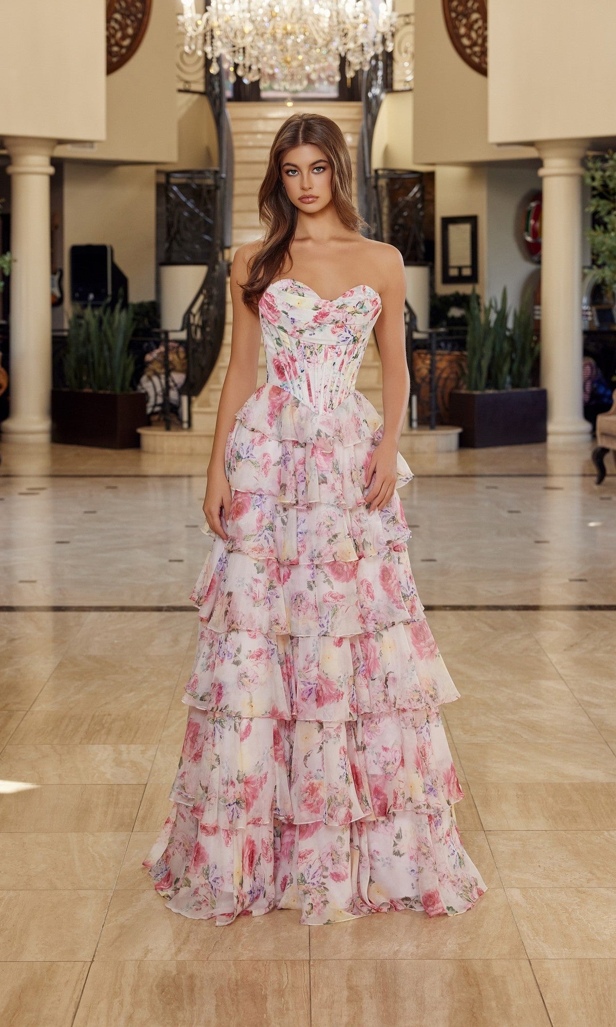 A woman stands indoors wearing the Nox Anabel Q1779 Long Prom Dress in Blush Print, a strapless corset style with pink floral pattern and tiered chiffon ruffles. Chandelier and staircase are in the background.